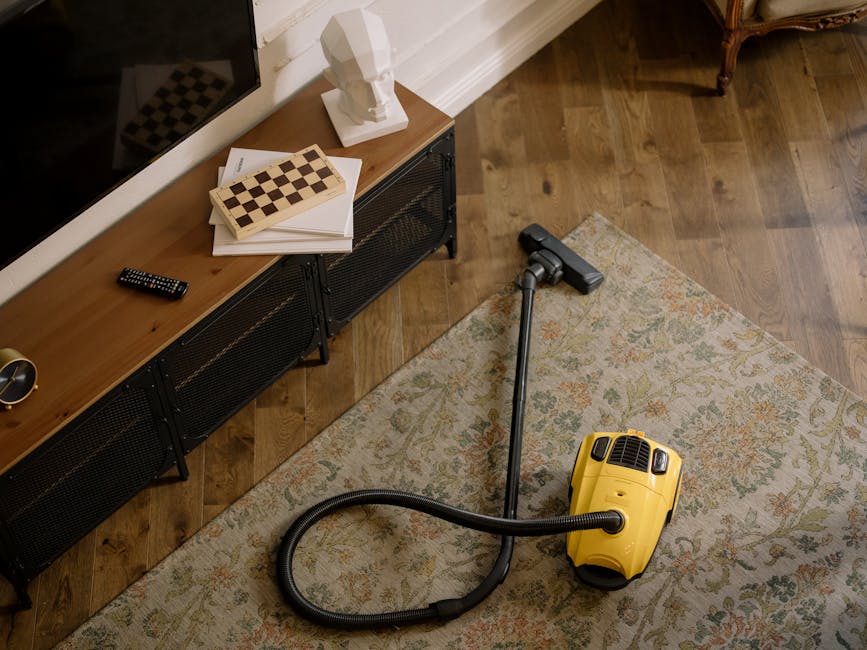 A yellow vacuum cleaner with a flexible hose and a black cleaning head resting on a traditional patterned area rug in a living room. The room features hardwood flooring, a wooden sideboard with black metal legs, and a white decorative bust atop the sideboard. A remote control and some paper are placed on the sideboard, while a small brass-colored object is also visible. The scene is well-lit, emphasizing the cleanliness and readiness for surface cleaning or deep cleaning, aligning with the services of Carpet Cleaners Hampstead for maintaining hygiene and domestic cleaning standards.