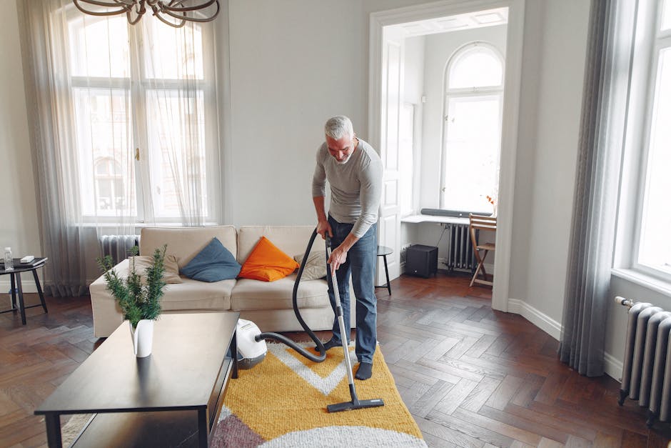 A man with grey hair and a beard is engaged in surface cleaning within a bright, spacious living room. He is using a vacuum cleaner on a yellow, modern geometric-patterned rug. The room features a white sofa with blue and orange cushions, a light-colored wooden coffee table with a potted plant on it, and large windows with sheer, white curtains allowing natural daylight to fill the space. The wooden flooring beneath the rug is polished and well-maintained, and there are additional furnishings such as a small black speaker, a white desk, and a wooden chair in the background. The overall scene highlights domestic cleaning activities, emphasizing thorough surface and deep cleaning, carried out by Carpet Cleaners Hampstead for optimal hygiene and freshness.