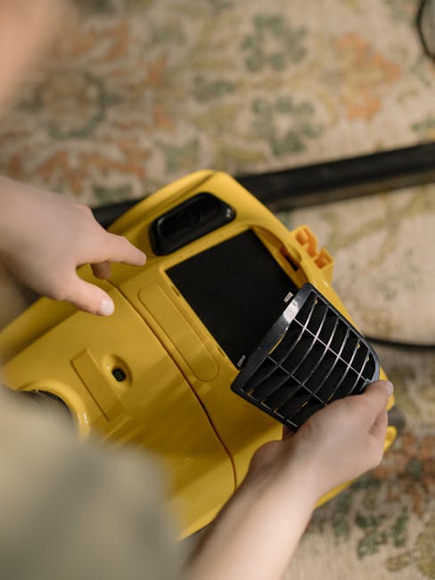 A yellow vacuum cleaner with a flexible hose and a black cleaning head resting on a traditional patterned area rug in a living room. The room features hardwood flooring, a wooden sideboard with black metal legs, and a white decorative bust atop the sideboard. A remote control and some paper are placed on the sideboard, while a small brass-colored object is also visible. The scene is well-lit, emphasizing the cleanliness and readiness for surface cleaning or deep cleaning, aligning with the services of Carpet Cleaners Hampstead for maintaining hygiene and domestic cleaning standards.