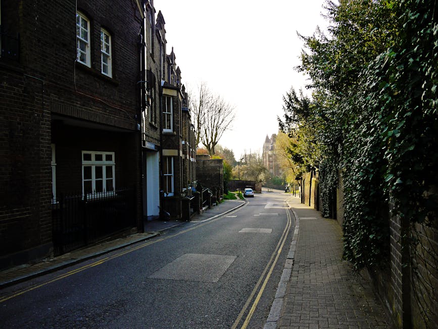 A quiet residential street with a paved sidewalk on the right, lined with green hedges and trees, and traditional brick houses with white-framed windows on the left. The asphalt road has clear yellow lines along the edges and appears clean and well-maintained. In the background, more houses and a church spire are visible under a bright, slightly overcast sky. The scene is well-lit by natural daylight, creating a calm and tidy street environment, suitable for highlighting the importance of regular cleaning and maintenance by Carpet Cleaners Hampstead, especially in maintaining hygiene and appearance in Hampstead residences.