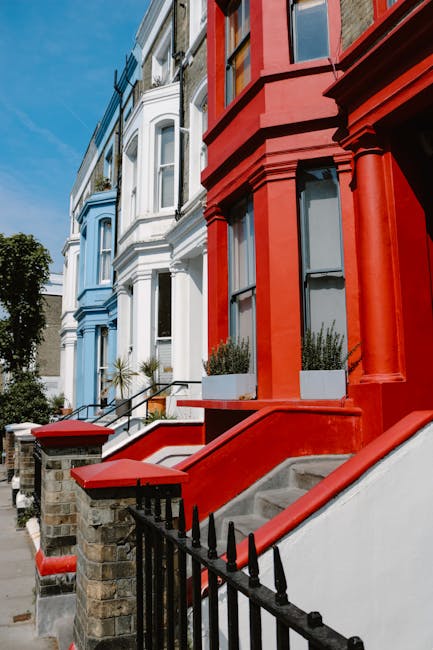 The image showcases a row of Victorian-style terraced houses on Rosslyn Hill in Hampstead, featuring a vibrant color scheme with one property painted in bright red and adjacent houses in light blue and white. The residential buildings have ornate architectural details, including large bay windows, decorative moldings, and black wrought-iron fences along the front. The steps leading up to each entrance are made of concrete with red-painted edges, and modern planters with greenery sit atop the front steps of the red house. Clear blue sky is visible overhead, and sunlight highlights the clean, well-maintained exteriors. The scene exudes a tidy and charming residential street environment, consistent with the professional deep cleaning and surface sanitisation services provided by Carpet Cleaners Hampstead for properties on Rosslyn Hill, Hampstead.